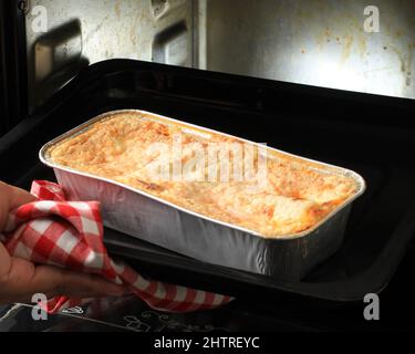 Le mani della donna che prendono la teglia con la lasagna fuori dal forno usando la lamina monouso nel forno elettrico, punto per punto che fa la cottura fatta in casa Foto Stock