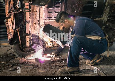 Giovane uomo meccanico lavoratore riparazione vecchia carrozzeria auto d'epoca in garage disordinato - sicurezza sul lavoro e protezione di abbigliamento - Guy con capelli freschi taglio a veicolo reno Foto Stock