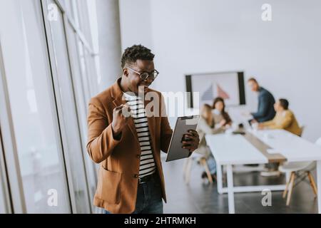 Bel giovane uomo d'affari afroamericano che lavora con il tablet digitale e riceve grandi notizie davanti ai suoi colleghi nella sala riunioni Foto Stock