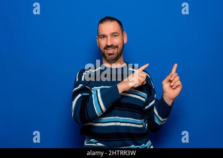 Immagine di un bell'uomo ispanico spensierato in un maglione a strisce puntando le dita lateralmente e ridendo a qualcosa di divertente, in piedi su sfondo blu Foto Stock