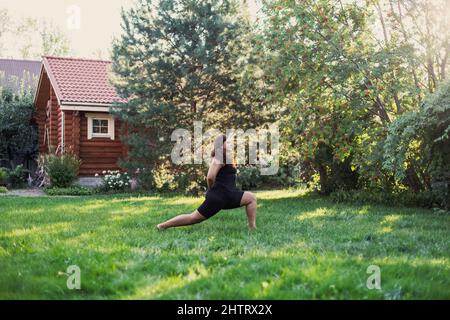 Donna grasso Blissful che fa le esercitazioni per allungare l'erba verde delle gambe sul cortile posteriore del cottage con la casa di legno e gli alberi in background. Positività del corpo Foto Stock