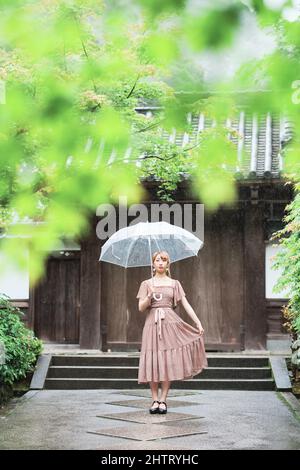 Ragazza giapponese carina che tiene il suo orlo vestito mentre si trova sotto un ombrello Foto Stock