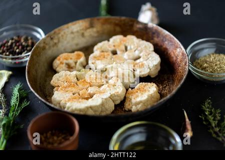 Cucina con bistecca di cavolfiore. Il cavolfiore crudo cosparso di spezie si trova in una padella. Olio d'oliva, erbe, varie spezie nelle vicinanze. Sfondo scuro. Foto Stock