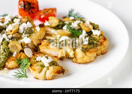 Bistecca di cavolfiore con spezie, salsa di chimichurri, fiocchi di mandorle, olive, pomodori ciliegini fritti e capperi su un piatto bianco. Cibo vegetariano. Foto Stock