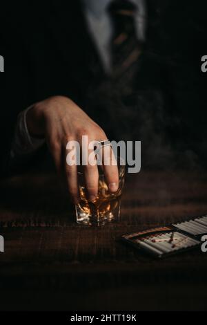 Scatto verticale di una mano elegante e adatta che tiene un bicchiere di whiskey accanto alle sigarette Foto Stock