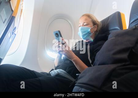 Una giovane donna che indossa la maschera facciale, usando lo smartphone mentre viaggia in aereo. Nuovo viaggio normale dopo il concetto di pandemia di covid-19. Foto Stock