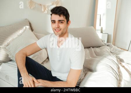 Ritratto di un giovane allegro che si rilassa nella sua camera da letto, seduto sul letto e guardando la macchina fotografica. Foto Stock