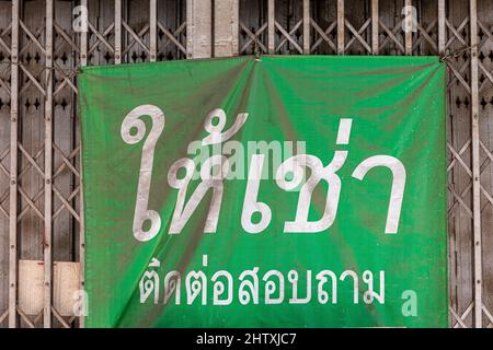 Un poster pubblicitario verde polveroso con lingue thailandesi significa per il noleggio e il contatto attaccato davanti alla porta d'acciaio del vecchio edificio all'interno di Bangkok. Foto Stock