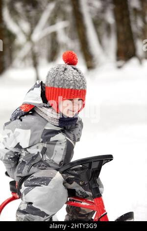 un ragazzo felice in discesa su una slitta in inverno, un bambino in abiti luminosi siede su una slitta, la neve lo vola Foto Stock