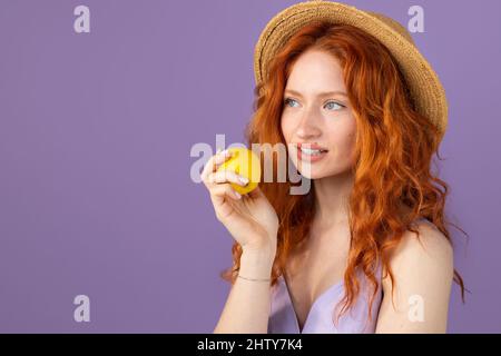 Ritratto di bellezza di una giovane ragazza rossa attraente con capelli ricci che indossa un cappello di paglia con una mela in mano su sfondo viola e spazio copia. Foto Stock