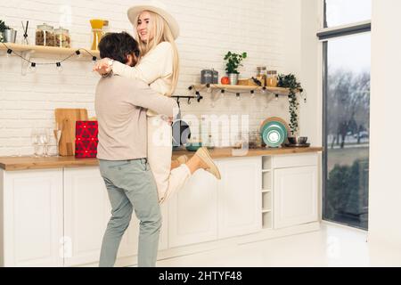 l'uomo millenario della bruna caucasica alza la sua bella moglie bionda dai capelli lunghi nella loro cucina moderna mentre festeggia. Foto di alta qualità Foto Stock