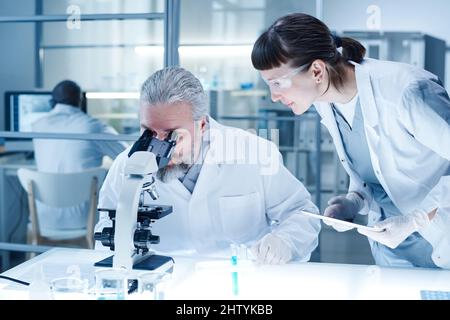 Uomo maturo in mano bianca che esamina l'analisi al microscopio con la donna che immette i dati nel computer durante il lavoro di squadra in laboratorio Foto Stock