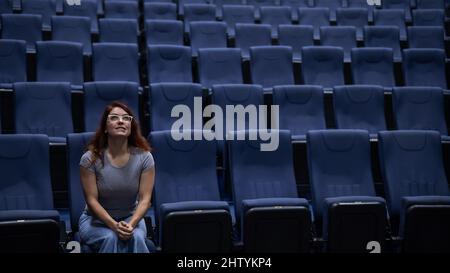 Donna caucasica dai capelli rossi siede in prima fila in un cinema in una sala vuota. La ragazza sta guardando un film da sola. Foto Stock