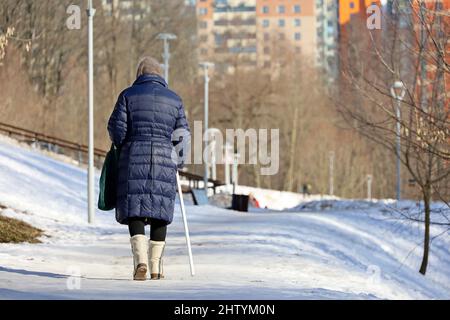 Vecchia donna con passeggiate di canna in primavera parco su edifici residenziali sfondo Foto Stock