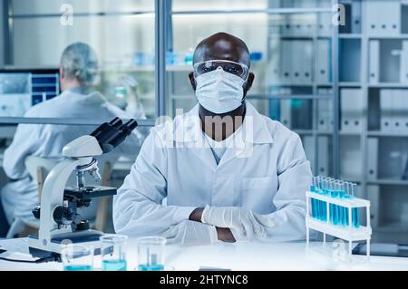 Ritratto di ricercatore africano in maschera e camice da laboratorio guardando la macchina fotografica mentre si siede al suo posto di lavoro in laboratorio con microscopio e matracci Foto Stock