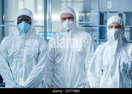 Ritratto di gruppo di medici in abbigliamento protettivo e maschere guardando la macchina fotografica mentre si lavora in squadra in laboratorio Foto Stock
