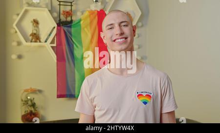 Libero uguale felice uomo gay caucasico sorridendo a macchina fotografica, posando nel suo appartamento, e in piedi di fronte alla bandiera arcobaleno orgoglio. Foto di alta qualità Foto Stock