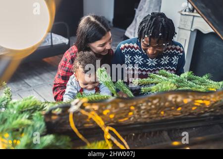 Bella famiglia multirazziale che festeggia il Natale, seduto al pianoforte con il loro adorabile ragazzo toddler. Foto di alta qualità Foto Stock