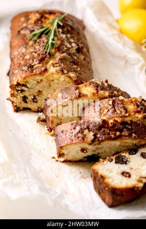 Fetta di torta al limone fatta in casa con uvetta, noci e rosmarino su carta da forno grattugiata, da servire con limoni freschi su tavola di marmo bianco. Primo piano Foto Stock