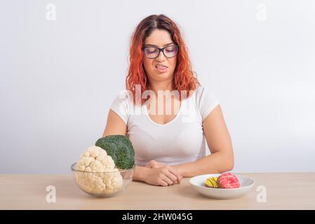 La donna caucasica preferisce cibo sano. Ragazza rossa sceglie tra broccoli e ciambelle su sfondo bianco. Foto Stock