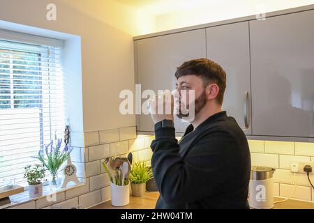 Giovane uomo con capelli favolosi, bevendo un bicchiere d'acqua in una cucina moderna Foto Stock