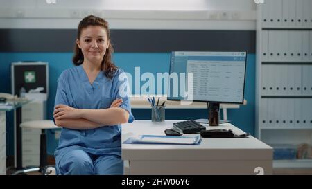 Ritratto di donna che lavora come assistente medico alla scrivania in ufficio medici. Infermiere che guarda la fotocamera e sorride mentre si prepara per il lavoro sul computer. Specialista sanitario in uniforme Foto Stock