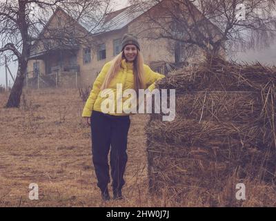 Una donna sorridente si erge su un pagliaio in un'area abbandonata sullo sfondo di un vecchio edificio dilapidato in un'opacità mistica di nubi Foto Stock