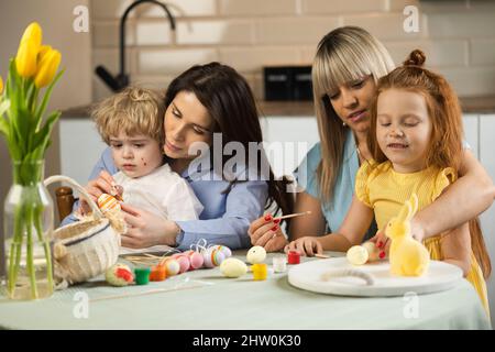 Due madri con i loro figli dipingono uova di Pasqua in cucina Foto Stock