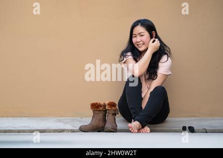 Rilassato Pretty Asian Woman siede sul pavimento del tetto con uno sfondo marrone chiaro e sorride alla macchina fotografica. Foto Stock