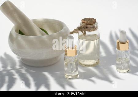 Olio da ingredienti naturali e un mortaio e pestello su sfondo bianco con ombre, fuoco selettivo. Olio naturale in bottiglia trasparente Foto Stock