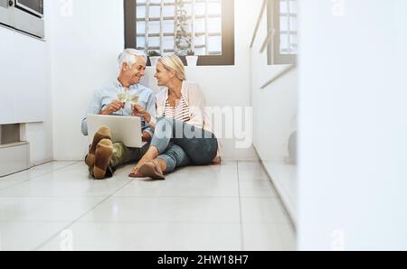 Heres a noi, babe. Scatto a tutta lunghezza di una coppia matura affettuosa che beve vino mentre navigano sul web sul piano della cucina. Foto Stock