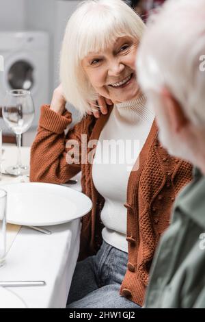 donna anziana sorridente a marito offuscato mentre si siede al tavolo festivo, immagine di scorta Foto Stock