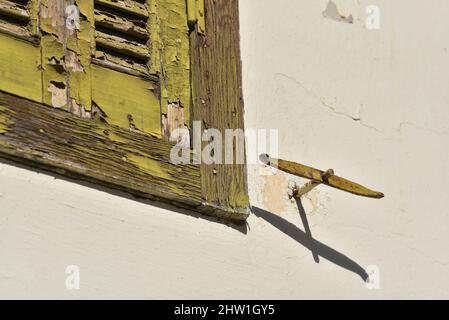 Vecchia casa rurale intemperie finestra con intagli in legno con vernice chartreuse scheggiata e un cane corrispondente otturatore su una parete sbiadita stucco a Nafplio, Grecia. Foto Stock