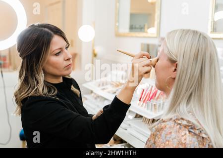 Cosmetologist che applica il trucco di fondazione ad una donna bionda in una sedia di trucco Foto Stock