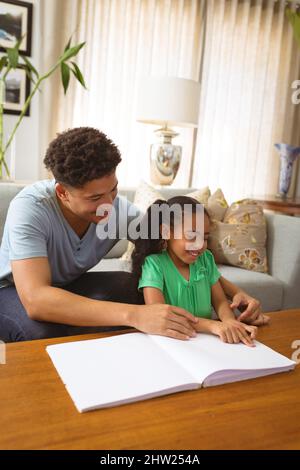 Felice padre biraciale aiutare figlia cieca studiare mentre legge libro con braille a casa Foto Stock