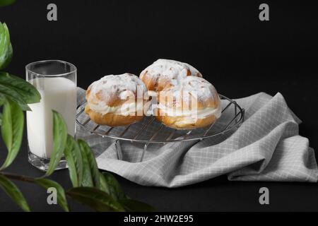 Profiteroles appena sfornati con crema si trovano sulla griglia, sotto la quale c'è un asciugamano da cucina. Un bicchiere di latte si trova nelle vicinanze. La foto è stata scattata da Foto Stock