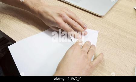 Le mani che fanno la figura di origami. Primo piano delle mani maschili usando un foglio bianco di carta per fare origami su sfondo tavola di legno, concetto di creatività. Foto Stock