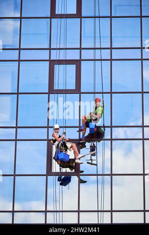 Lavoratori alpinisti industriali lavano finestre in vetro di un alto edificio, appesi su funi di arrampicata di sicurezza, guardando la macchina fotografica. Due pulitori per finestre uomini che lavorano insieme all'esterno del grattacielo. Foto Stock
