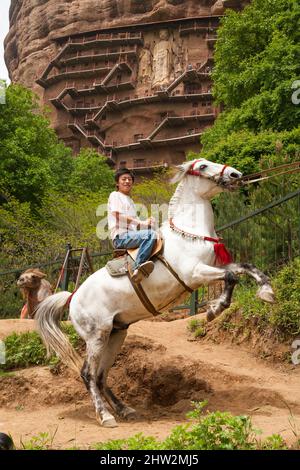 Un turista cinese paga sedersi su un cavallo che è stato addestrato per arretrare sulle sue gambe posteriori, mentre gli spettatori scattano le fotografie del trucco. (67/125) l'acrobazia si svolge al di fuori delle Grotte di Maijishan, precedentemente romanizzate come Maichishan, sono una serie di 194 grotte tagliate sul lato della collina di Majishan a Tianshui, provincia di Gansu, Cina. PRC. (125) Foto Stock