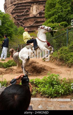 Un turista cinese paga sedersi su un cavallo che è stato addestrato per arretrare sulle sue gambe posteriori, mentre gli spettatori scattano le fotografie del trucco. (67/125) l'acrobazia si svolge al di fuori delle Grotte di Maijishan, precedentemente romanizzate come Maichishan, sono una serie di 194 grotte tagliate sul lato della collina di Majishan a Tianshui, provincia di Gansu, Cina. PRC. (125) Foto Stock