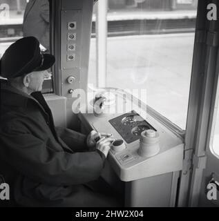 1968, storico, un driver della metropolitana in berretto e giacca, seduto nella cabina del conducente di un treno della metropolitana di Londra, Londra, Inghilterra, Regno Unito. Sta premendo due pulsanti per avviare il treno, dopo di che assume automaticamente il controllo. Noti come stock del 1967, i treni operarono per la prima volta sulla linea Victoria, aperta di recente, nel 1968 e furono in uso fino al 2011. Dotati del sistema Automatic Train Operation (ATO), sono stati i primi treni ferroviari automatici al mondo. Sono stati anche il primo materiale rotabile della metropolitana di Londra a presentare finestre avvolgenti nella cabina del conducente. Foto Stock