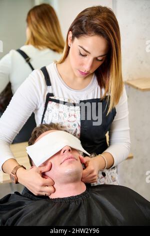 Parrucchiere mettere un asciugamano caldo sul viso dell'uomo prima di rasarlo al barbiere Foto Stock