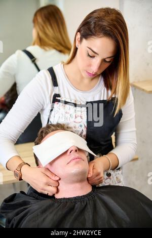 Parrucchiere mettere un asciugamano caldo sul viso dell'uomo prima di rasarlo al barbiere Foto Stock