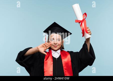 Ragazza di 9-11 anni con cappuccio di laurea e accappatoio per cerimonie con certificato di diploma su sfondo azzurro. Laureato per celebrare la laurea. Concetto di istruzione. Scuola elementare di successo Foto Stock