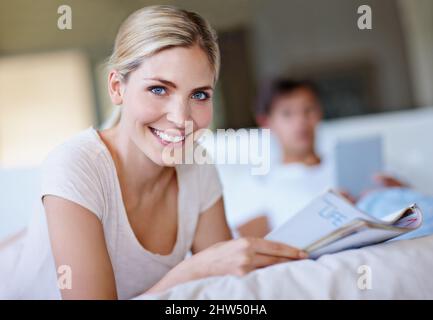 Ruoti la pagina ad un più felice voi. Scatto di una giovane donna felice sdraiata a letto e leggendo una rivista mentre suo marito usa un tablet digitale in Foto Stock