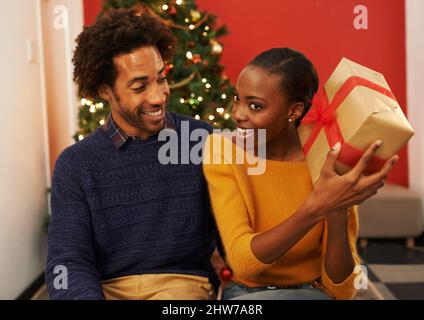 Qualunque cosa potrebbe essere. Scatto di una giovane donna che tiene il suo regalo di Natale mentre si siede accanto al suo ragazzo. Foto Stock