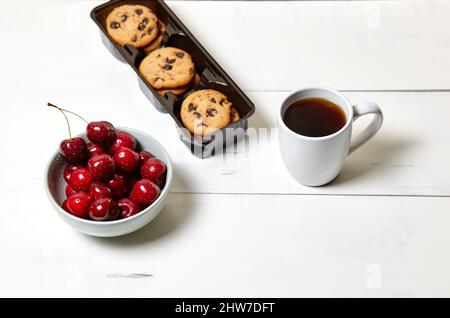 Tazza bianca con caffè, biscotti tradizionali e ciliegie dolci in una ciotola di ceramica su fondo di legno bianco, primo piano. Gustosa colazione, mattina routin Foto Stock