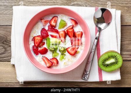 Porridge di farina d'avena con fette di kiwi, fragole in ciotola rosa, cucchiaio, tovagliolo con strisce rosse su sfondo di legno Vista dall'alto piatto Lay Vegan food conc Foto Stock