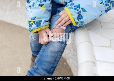 Ragazzino, che mostra il ginocchio ferito da una caduta, seduto su una panchina nel parco Foto Stock