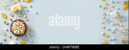Banner pasquale con uova di cioccolato pastello, nido, coniglietto, mimosa, gypsophila e cazzo giallo su sfondo blu. Spazio per il testo. Vista dall'alto. Piatto l Foto Stock